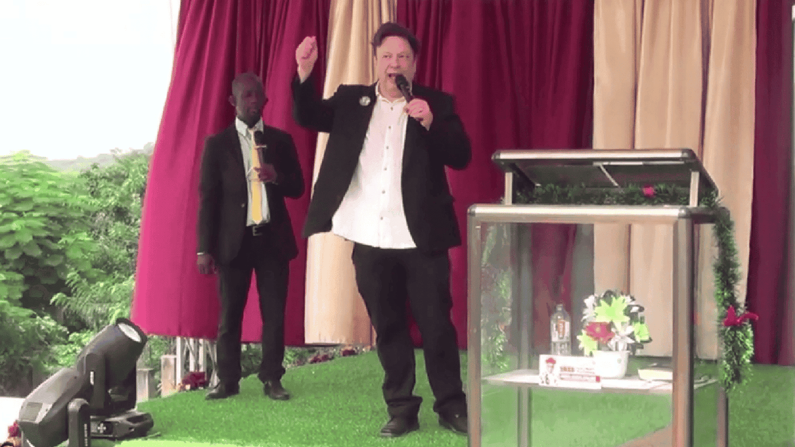 Man speaking at an outdoor event with two men in the background.