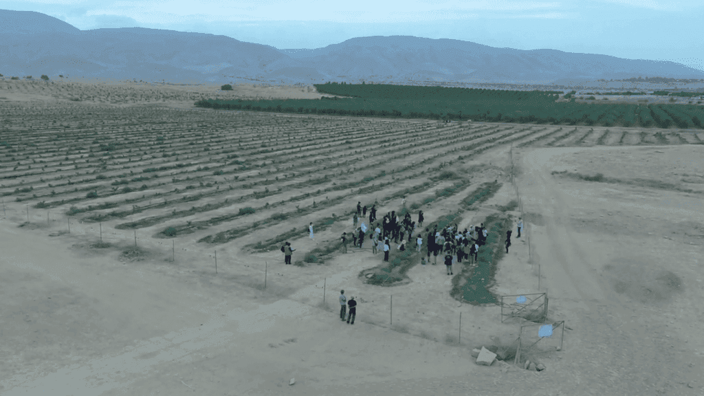 Group of people gathered in a vast, arid agricultural landscape with mountains in the background.